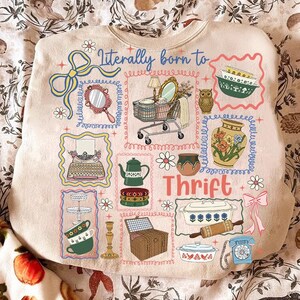 May include: Beige sweatshirt with a colorful graphic design featuring various vintage items like a mirror, shopping cart, and teapot. The text "Literally born to Thrift" is displayed in blue script, with additional decorative elements.