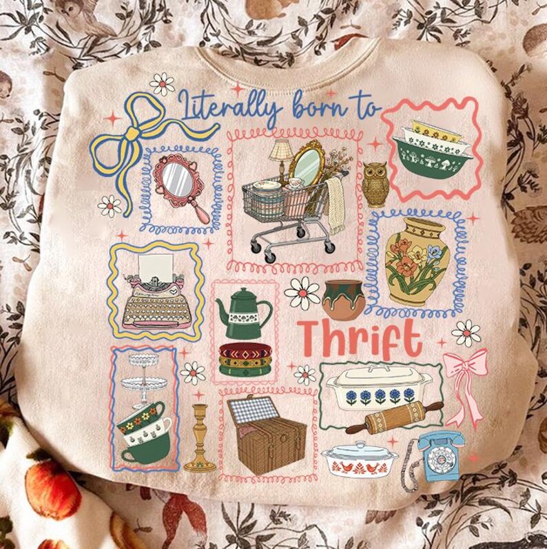 May include: Beige sweatshirt with a colorful graphic design featuring various vintage items like a mirror, shopping cart, and teapot. The text "Literally born to Thrift" is displayed in blue script, with additional decorative elements.
