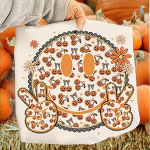 May include: A white decorative pillow featuring a smiling face design with orange eyes and a black lace border. The face is decorated with orange pumpkins and peace signs, and orange flowers. The background includes pumpkins.