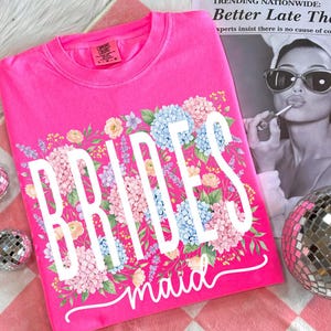 May include: A bright pink t-shirt with the words "BRIDESmaid" in white script, surrounded by a floral design of blue, pink, and yellow flowers. The shirt is a Comfort Colors brand.