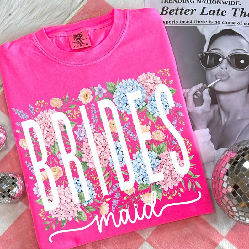 May include: A bright pink t-shirt with the words "BRIDESmaid" in white script, surrounded by a floral design of blue, pink, and yellow flowers. The shirt is a Comfort Colors brand.