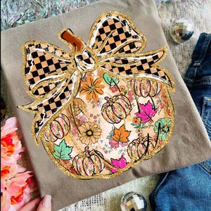 May include: A tan t-shirt with a pumpkin design. The pumpkin is decorated with flowers, pumpkins, and leaves in various colours. A gold glitter outline and a checkered bow complete the design.