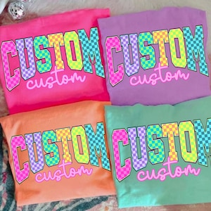 May include: Four colorful t-shirts in pink, purple, orange, and green. Each shirt features the word "CUSTOM" in a bold, checkered design, with the word "custom" below. The shirts are folded and arranged on a patterned surface.