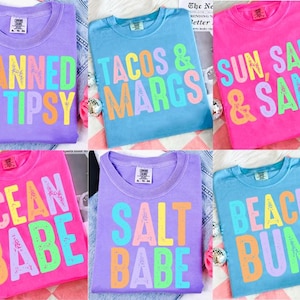 Könnte beinhalten: Eine Auswahl an bunten T-Shirts mit verschiedenen Sätzen in fetten, mehrfarbigen Buchstaben. Die Shirts sind in Lila, Blau und Pink gehalten. Sätze sind "TANNED & TIPSY", "TACOS & MARGS", "SUN, SALT & SAND", "OCEAN BABE", "SALT BABE" und "BEACH BUM".