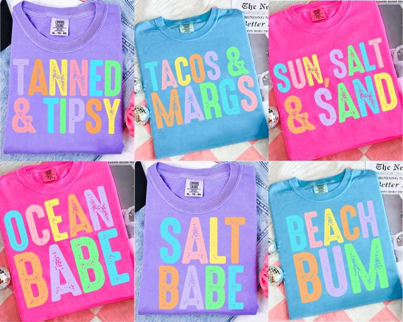 May include: A collection of colorful t-shirts with various phrases in bold, multi-colored letters. The shirts are in shades of purple, blue, and pink. Phrases include "TANNED & TIPSY", "TACOS & MARGS", "SUN, SALT & SAND", "OCEAN BABE", "SALT BABE", and "BEACH BUM".