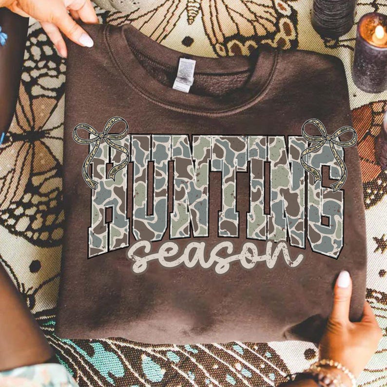 May include: A brown sweatshirt with the words "HUNTING season" in a camouflage pattern. The sweatshirt is being held up by a person.