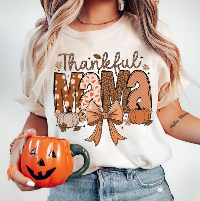 Op de afbeelding: Een wit T-shirt met een bruine en oranje afbeelding met de tekst "Thankful Mama" met een strik en pompoenen.