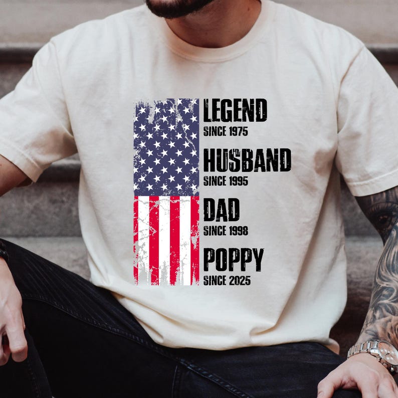 K&ouml;nnte beinhalten: Cremefarbenes T-Shirt mit einem verblassten Design der amerikanischen Flagge. Die Flagge ist in zwei Teile geteilt, wobei die obere H&auml;lfte die Sterne und die untere H&auml;lfte die Streifen zeigt. Text: "LEGEND SINCE 1975", "HUSBAND SINCE 1995", "DAD SINCE 1998" und "POPPY SINCE 2025".