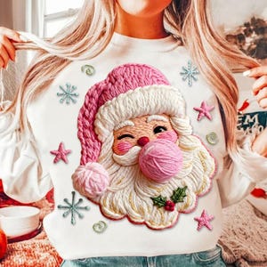 May include: White long-sleeved sweatshirt featuring a Santa Claus design. The Santa Claus face is embroidered with pink, white, and cream yarn. Pink stars and snowflakes are embroidered around the Santa Claus face. A festive holiday-themed sweatshirt.