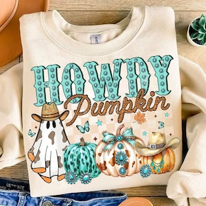 May include: A cream-colored sweatshirt with the text "Howdy Pumpkin" in turquoise and brown lettering. The design features a ghost wearing a cowboy hat, turquoise and brown pumpkins, and a turquoise and brown bow.