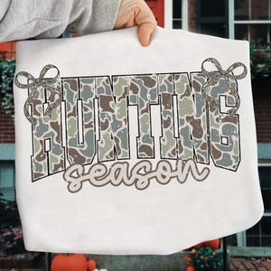 May include: White sweatshirt featuring the text "HUNTING season" in a camouflage pattern. The word "HUNTING" is in large, block letters, and "season" is in a cursive font. Decorative bows are on each side of the word "HUNTING".