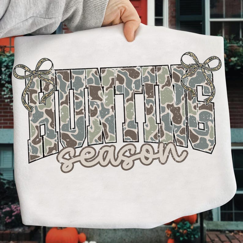 May include: White sweatshirt featuring the text "HUNTING season" in a camouflage pattern. The word "HUNTING" is in large, block letters, and "season" is in a cursive font. Decorative bows are on each side of the word "HUNTING".