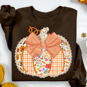 K&ouml;nnte beinhalten: Ein braunes Sweatshirt mit einem K&uuml;rbisdesign. Der K&uuml;rbis besteht aus orangefarbenem Vichy-Karo, floralen Mustern und einer gro&szlig;en orangefarbenen Schleife. Das Design ist mit einer wei&szlig;en Spitzenborte umrandet und schafft ein festliches Herbstthema.