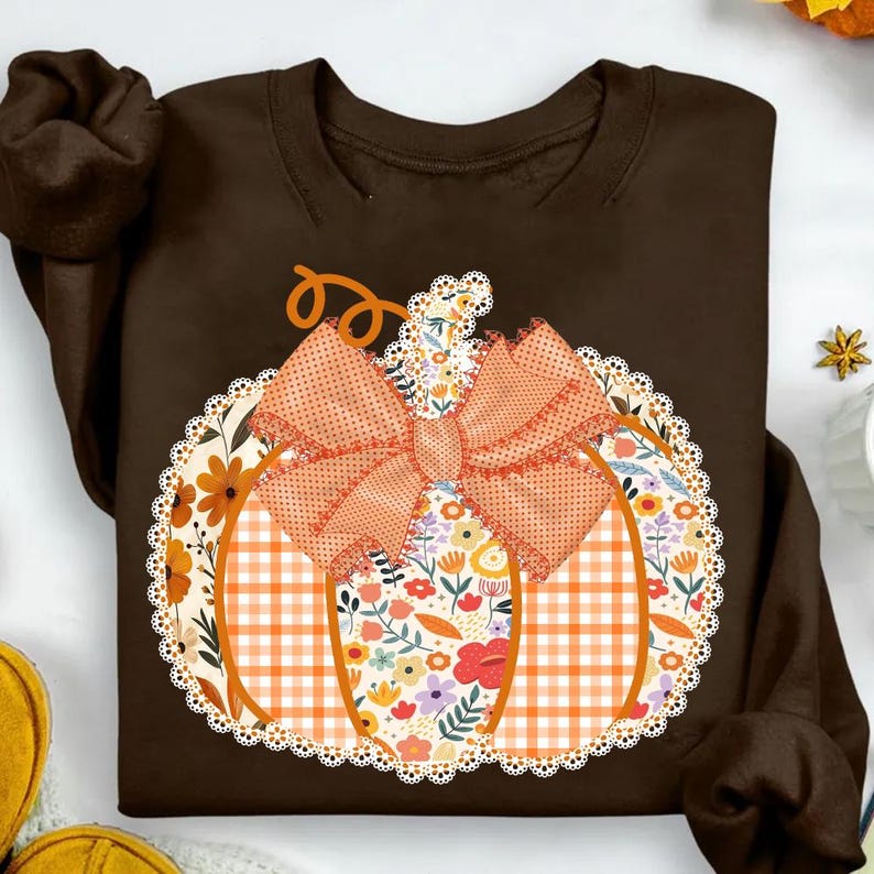 K&ouml;nnte beinhalten: Ein braunes Sweatshirt mit einem K&uuml;rbisdesign. Der K&uuml;rbis besteht aus orangefarbenem Vichy-Karo, floralen Mustern und einer gro&szlig;en orangefarbenen Schleife. Das Design ist mit einer wei&szlig;en Spitzenborte umrandet und schafft ein festliches Herbstthema.