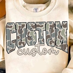 Könnte beinhalten: Cremefarbenes Sweatshirt mit dem Wort "CUSTOM" in großen Buchstaben mit Tarnmuster, schwarz umrandet. Darunter steht das Wort "custom" in Schreibschrift. Das Sweatshirt hat einen Rundhalsausschnitt.