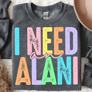 May include: Dark gray sweatshirt with the text "I NEED an ALANI" in colorful, distressed lettering. The text is in various colors, including blue, pink, yellow, orange, green, and purple. The sweatshirt has a crew neck.