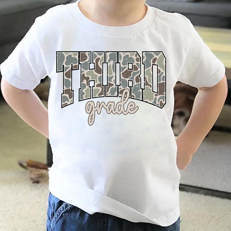 May include: White t-shirt with the words "THIRD grade" in a camouflage pattern. The word "grade" is in a cursive font. The shirt is made of a soft material and is suitable for children.