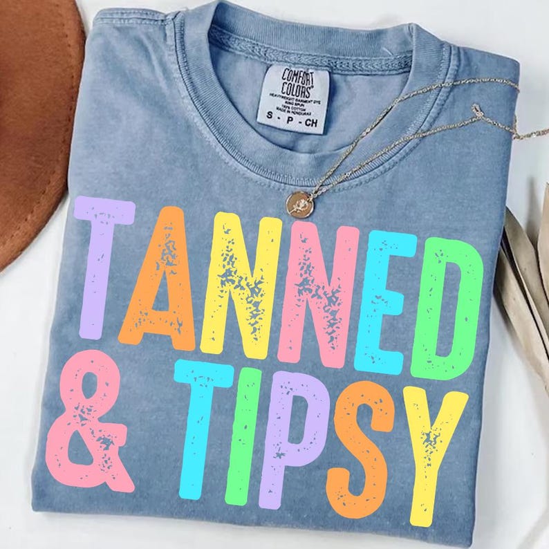 May include: A blue Comfort Colors t-shirt with the words "TANNED & TIPSY" in colorful, distressed lettering. A gold necklace with a pendant rests on the shirt. The shirt is size small.