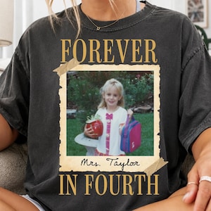 May include: A charcoal gray short-sleeve t-shirt featuring a vintage-style graphic print. The design includes the text FOREVER IN FOURTH in gold, framing a photograph of a child holding a red apple and a pink lunchbox, with Mrs. Taylor written below.