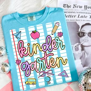 Peut inclure: T-shirt bleu clair avec un motif color&eacute; "Kindergarten". Le motif comprend un avion en papier, une pomme, un crayon, des fleurs, un bus scolaire, un globe et un n&oelig;ud. Le t-shirt a un col rond et des manches courtes.
