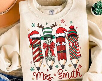 Puede incluir: Sudadera color crema con un diseño festivo de lápices. Cada lápiz está decorado con elementos navideños como gorros de Papá Noel, bufandas y cuernos de reno. El texto "Mrs. Smith" está impreso en rojo debajo de los lápices, rodeado de copos de nieve.