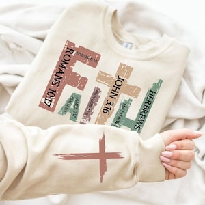 Puede incluir: Una sudadera beige con un diseño gráfico de cruces y versículos bíblicos, incluyendo "Romanos 10:17", "Juan 3:16", "Hebreos", "Efesios 2:8", "Proverbios 3:5-6", "Santiago 2:17" y "Salmo 37:5".