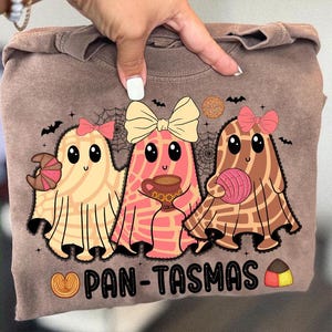May include: Brown sweatshirt featuring a graphic of three ghost-like characters, each adorned with a bow and holding various treats. The text "PAN-TASMAS" is displayed below the characters. The design includes bats, spiderwebs, and decorative elements.