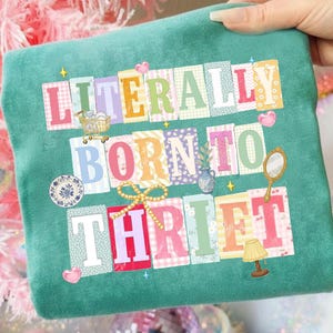 Op de afbeelding: Tealkleurige sweatshirt met de tekst "LITERALLY BORN TO THRIFT" in kleurrijke, patroonletters. Decoratieve elementen zijn een winkelwagentje, een vaas, een spiegel, een lamp en harten.