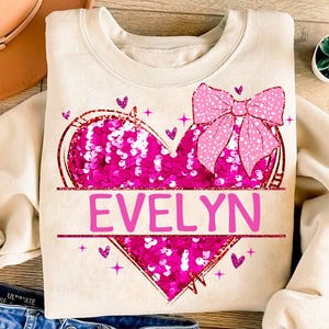 May include: A cream-colored sweatshirt with a pink sequined heart and a pink bow. The heart has the name "EVELYN" written across it.
