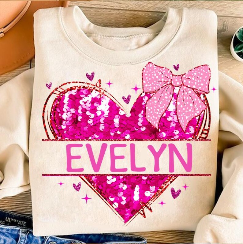 May include: A cream-colored sweatshirt with a pink sequined heart and a pink bow. The heart has the name "EVELYN" written across it.