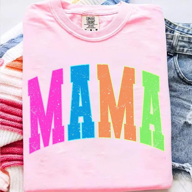 Może przedstawiać: Jasnor&oacute;żowa koszulka z napisem "MAMA" dużymi, kolorowymi, zniszczonymi literami. Litery są w odcieniach r&oacute;żu, niebieskiego, pomarańczowego i zielonego. Koszulka jest złożona, z widoczną metką przy dekolcie.