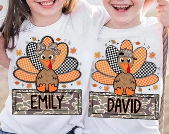 May include: White t-shirts featuring cartoon turkeys with orange and black checkered tail feathers. One turkey has a bow, the other a hat. Each shirt has a nameplate with the names "EMILY" and "DAVID" in a camouflage design, perfect for Thanksgiving.