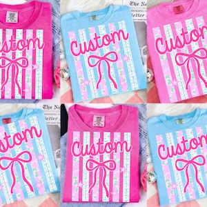 May include: A collection of pink and blue t-shirts with a custom design. Each shirt features the word "Custom" in a cursive font, a bow, and vertical stripes with star accents. The shirts are folded and displayed.
