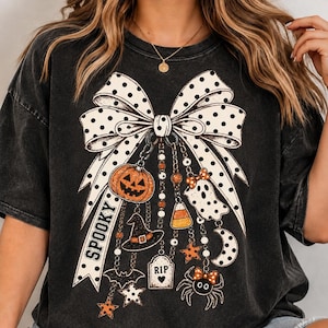 May include: A charcoal black t-shirt featuring a large white polka dot bow graphic. Hanging from the bow are various Halloween-themed charms including a jack-o'-lantern, a witch hat, a tombstone labeled RIP, a ghost, a spider, and stars.