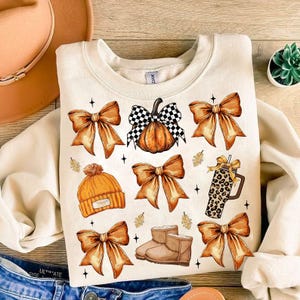 May include: Cream-colored sweatshirt featuring a fall-themed design. The design includes orange bows, a pumpkin with a checkered bow, a beanie, boots, and a leopard-print tumbler. The overall theme is autumn and seasonal.