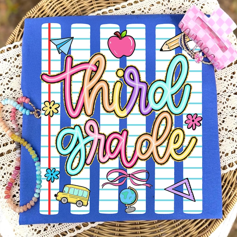 Peut inclure: T-shirt bleu avec un motif color&eacute; "troisi&egrave;me ann&eacute;e". Le design comprend un avion en papier, une pomme, un crayon, des fleurs, un bus scolaire, un n&oelig;ud, un globe et un triangle. Un collier de perles et une pince &agrave; cheveux rose sont &eacute;galement visibles.