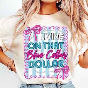 May include: A cream-colored t-shirt featuring a graphic design with the text "LIVING ON THAT Blue Collar DOLLAR". The design includes a light blue background with white stripes, pink bows, and a decorative border.