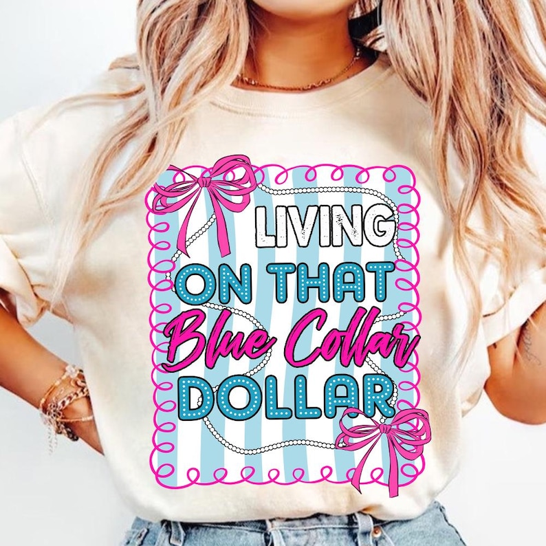 May include: A cream-colored t-shirt featuring a graphic design with the text "LIVING ON THAT Blue Collar DOLLAR". The design includes a light blue background with white stripes, pink bows, and a decorative border.
