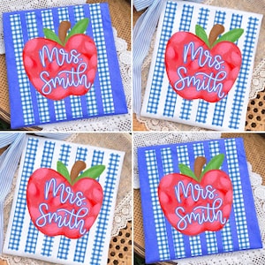 May include: Four square decorative towels with a red apple design and the text "Mrs. Smith". The apple has green leaves and a brown stem. The towels have a blue and white gingham pattern.
