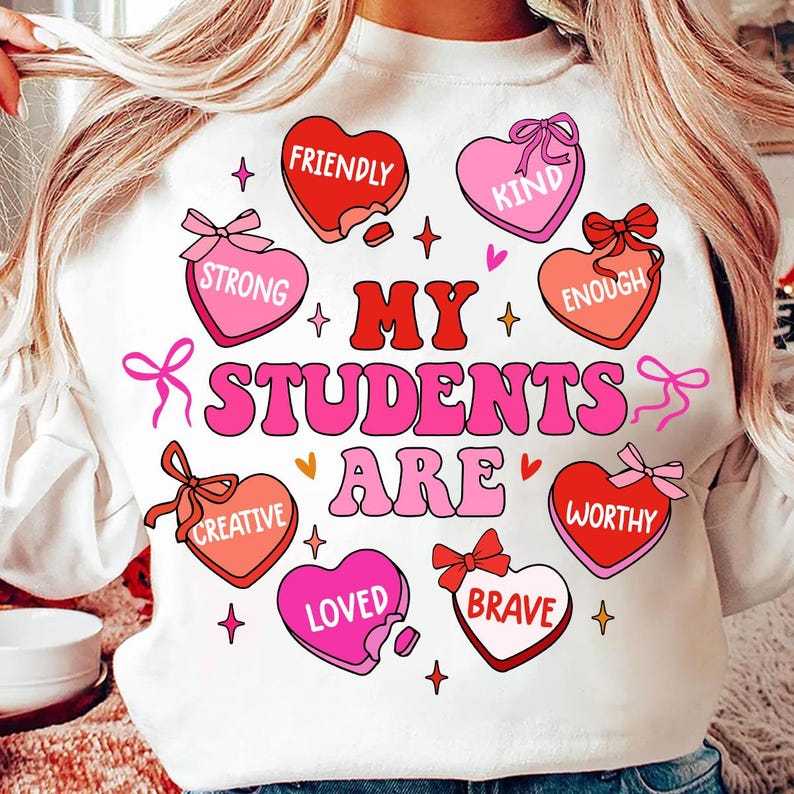 May include: White sweatshirt with a pink and red heart design. The hearts have words on them like "Friendly", "Kind", "Strong", "Enough", "Creative", "Loved", "Brave", and "Worthy". The text "My Students Are" is in the center of the design.