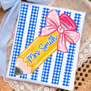 May include: A white and blue gingham patterned design with a yellow pencil graphic. The pencil has the name "Mrs. Smith" in blue script and is adorned with a pink bow. A pink eraser is attached to the pencil.