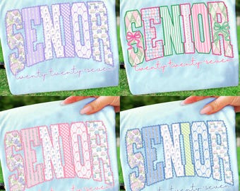 Puede incluir: Cuatro camisetas azul claro con la palabra "SENIOR" en diferentes combinaciones de colores. El texto "twenty twenty-seven" está bordado debajo de la palabra "SENIOR". Los diseños incluyen elementos florales, a rayas y estampados.