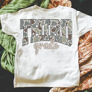 May include: White t-shirt featuring the words "THIRD grade" in a camouflage pattern. The word "grade" is in a cursive font. The shirt is made of a soft material and is suitable for children.