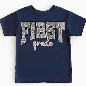 May include: Navy blue toddler t-shirt with the words "FIRST grade" printed on the front. The word "FIRST" is in large, block letters with a camouflage pattern. The word "grade" is in a cursive font.