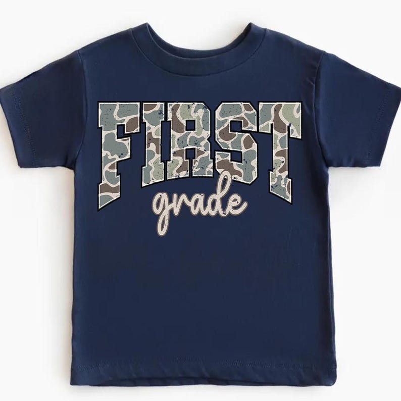 May include: Navy blue toddler t-shirt with the words "FIRST grade" printed on the front. The word "FIRST" is in large, block letters with a camouflage pattern. The word "grade" is in a cursive font.