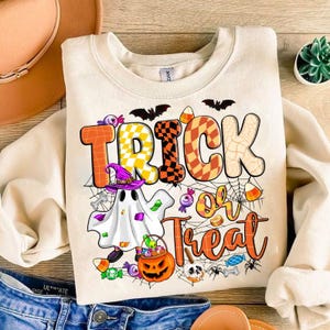 May include: A cream-colored sweatshirt with a graphic print of a ghost holding a bucket of candy. The graphic also includes the text "Trick or Treat" with a checkered pattern and spiderwebs.