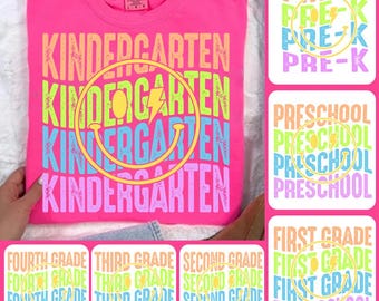 May include: A vibrant pink t-shirt with the word "KINDERGARTEN" in a rainbow gradient, featuring a smiley face with a lightning bolt. Additional images show "PRE-K", "PRESCHOOL", and "FIRST GRADE" designs in similar styles.