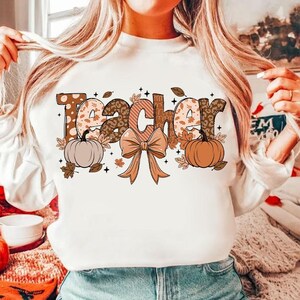 May include: White sweatshirt with the word "Teacher" in brown, orange, and white patterned letters. The letters are decorated with fall-themed elements like pumpkins, leaves, and a bow.