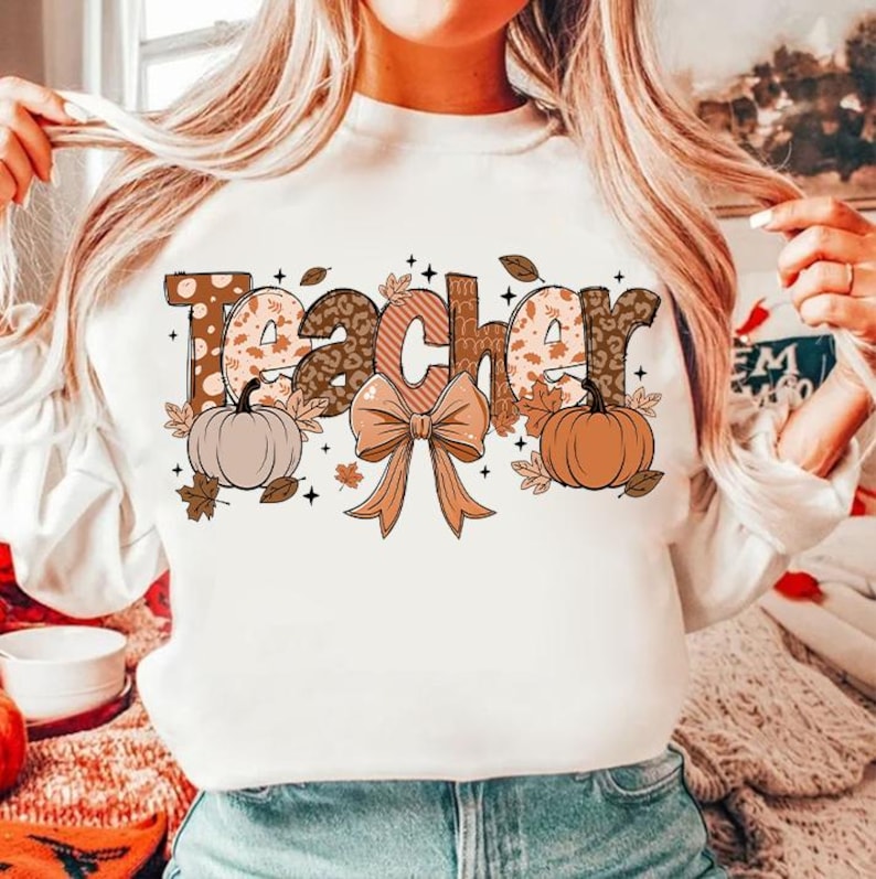May include: White sweatshirt with the word "Teacher" in brown, orange, and white patterned letters. The letters are decorated with fall-themed elements like pumpkins, leaves, and a bow.