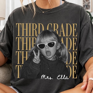 Op de afbeelding: Een antracietgrijs t-shirt met korte mouwen en de herhaalde gouden tekst THIRD GRADE. In het midden staat een zwart-witfoto van een kind met staartjes en een zonnebril die een vredesteken maakt, met de witte scripttekst Mrs. Ella eronder.